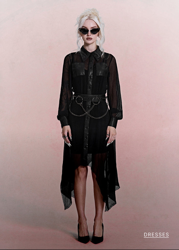 A model with platinum blonde hair and sunglasses wearing a black sheer long-sleeve shirt dress with a high-low hem, a matching black belt with a chain link detail, and black heels, standing against a solid pink studio background.